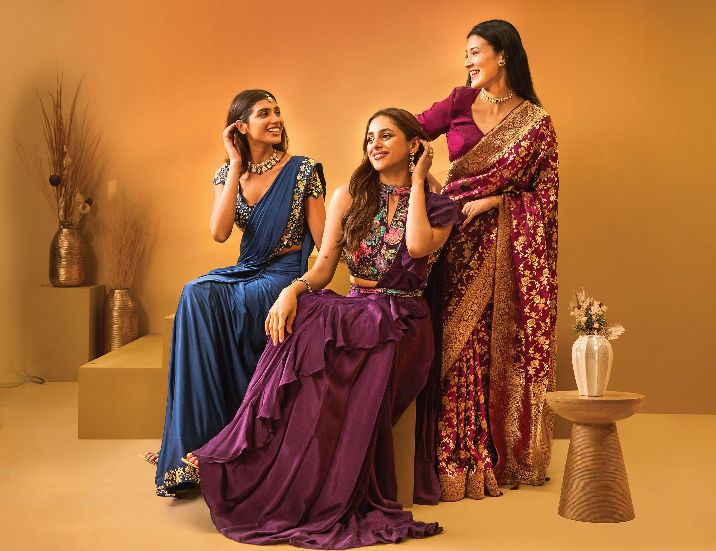 Three women wearing elegant Indian ethnic outfits including a sari and stylish lehengas in jewel tones, posing joyfully against a warm background